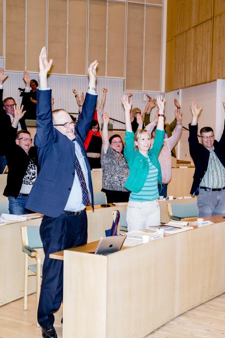 Nousevassa katsomossa olevat ihmiset ovat nousseet seisomaan ja nostaneet molemmat kädet ylös. Moni hymyilee. Pöydillä heidän edessään on läppäreitä, papereita ja kyniä.