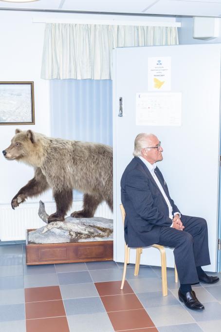 Vanhempi mies sinisessä puvussa istuu tuolilla metallisen oven edessä. Hänen takanaan seinän vieressä on täytetty karhu, joka katsoo eri suuntaan kuin mies.
