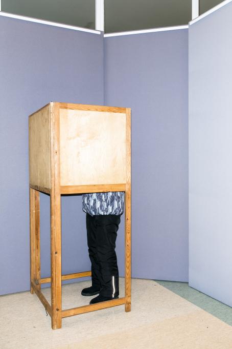 Puinen äänestyskoppi, jossa on joku sisällä. Hänestä näkyvät vain ulkohousuihin verhoutuneet jalat.
