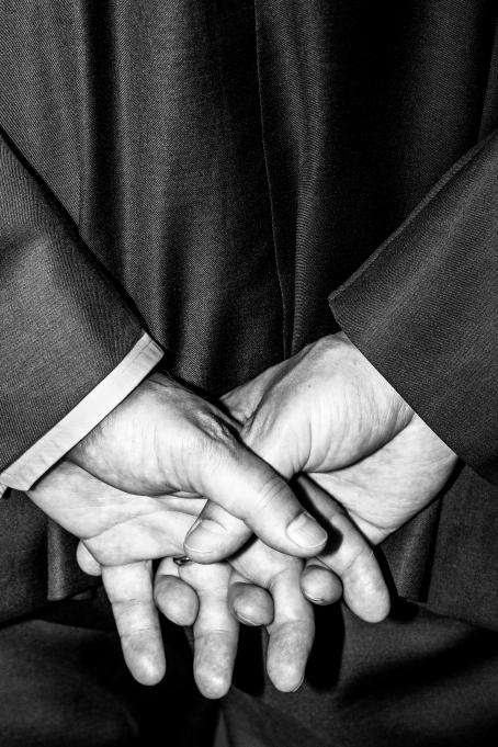 Sakari Piippo: Elinkeinoministeri Olli Rehn (kesk.) tiedotustilaisuudessa koskien Terrafame Oyn kaivostoimintaa Sotkamossa 18.8.2015, sarjasta Eräitä huomioita Suomen poliittisesta järjestelmästä, 2015–2019. Mustavalkoisessa kuvassa henkilön kädet. Hän pitää toisella kämmenellä kiinni toisesta selkänsä takana.