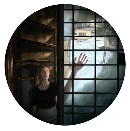 Pyöreässä kuvassa nainen seisoo ristikkomaisen oven vieressä silmät kiinni. Hänen vieressä olevassa ovessa/ikkunassa tms. näkyy katumaisemaa ja ihmisen käsivarsi vasten sen pintaa. 