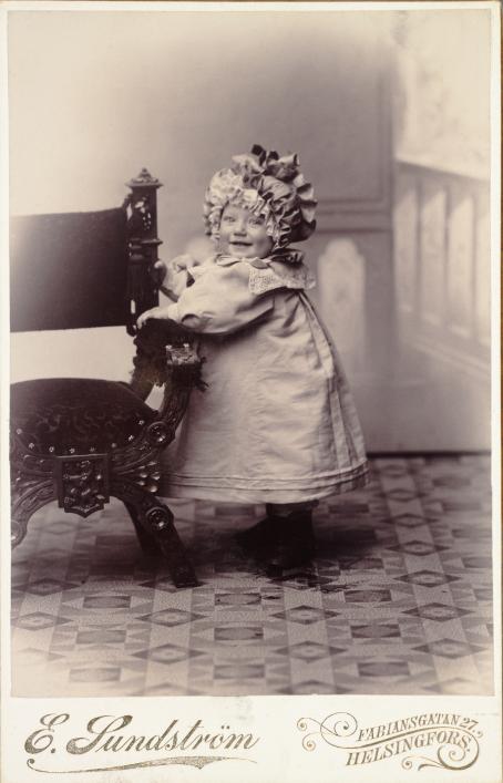 Mustavalkoisessa kuvassa pieni lapsi puettuna mekkoon sekä isohkoon, rypytettyyn hattuun seisoo ottaen tukea tuolista. Hän hymyilee ja katsoo kameraan.