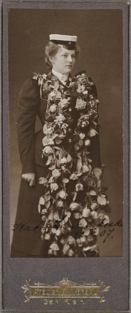 Mustavalkoisessa kuvassa nainen tummassa mekossa seisoo ylioppilaslakki päässään. Hänellä on kaulassaan iso, pitkä, lähes maahan asti ulottuva kukkaseppele. 
