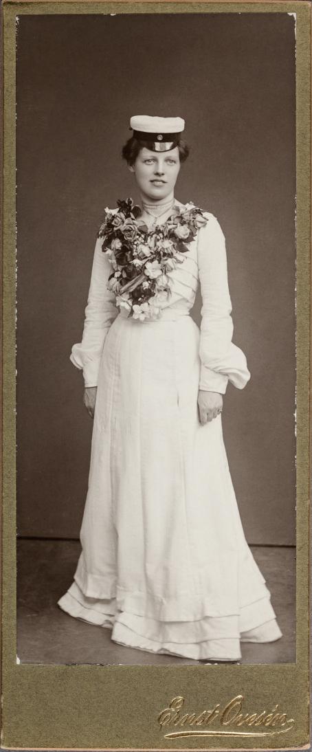 In a black and white photo a woman is standing dressed in a white, long dress. Bodice of the dress is full of flowers. She is wearing a Finnish student cap. 