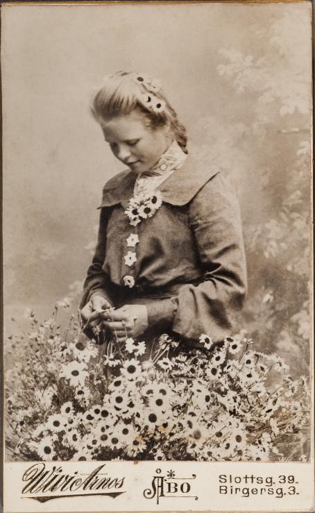 In a black and white photo a young woman is standing looking downward. There’s a bunch of flowers in front of her, and she is fiddling with one flower. There are flowers also in her jacket’s collar and in her hair. 