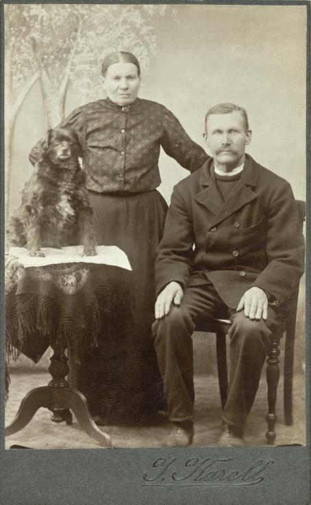 Mustavalkoisessa kuvassa mies, nainen ja koira. Mies istuu oikealla tuolilla ja koira istuu pöydällä vasemmalla puolella. Nainen seisoo heidän takanaan, pitäen toista kättä lanteillaan ja toista miehen tuolin päällä. 