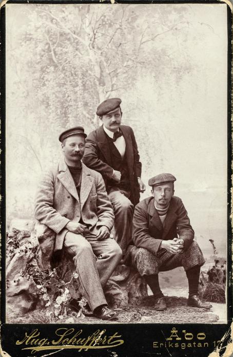 In a black and white photo there are three men sitting on different levels but close to each other. The background has a painted birch tree. 