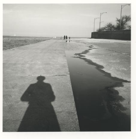 Vivian Maier, 1956. © Estate of Vivian Maier, Courtesy of Maloof Collection and Howard Greenberg Gallery, NY. Mustavalkoinen rantatienäkymä. Vasemmalla puolella vettä, edessä kävelytietä ja kaukana ihmisiä. Tiehen heijastuu ihmisen varjo.