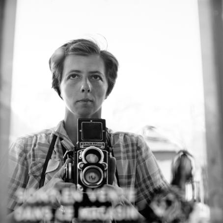 Vivian Maier, 1959. © Estate of Vivian Maier, Courtesy of Maloof Collection and Howard Greenberg Gallery, NY. Mustavalkoinen omakuva lyhythiuksisesta, ruutupaitaisesta naisesta. Hän pitelee Rolleiflex-kameraa. Kuvan alareunassa lukee jotain, mutta kirjaimet ovat epäselviä.