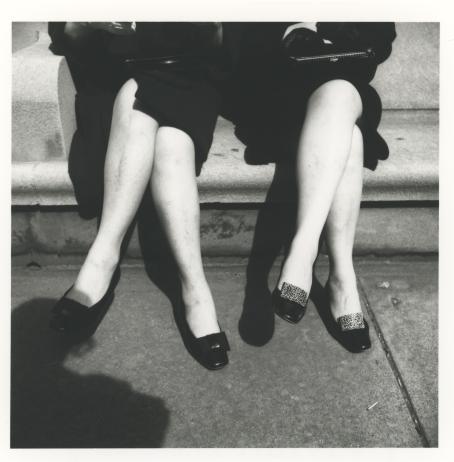Mustavalkoisessa kuvassa kahden naisen jalat. Naiset istuvat portaalla vierekkäin, heillä molemmilla on mustat takit ja mustat kengät, mutta sääret ovat paljaat. Molemmat ovat nostaneet vasemman jalan oikean päälle ristiin. Kuvaajan varjo näkyy kuvan vasemmassa alalaidassa. 