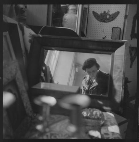 Vivian Maier, Chicagon alue, 1971. © Estate of Vivian Maier, Courtesy of Maloof Collection and Howard Greenberg Gallery, NY. Mustavalkoinen omakuva. Kuvan keskellä olevassa peilissä näkyy baskerihattuinen nainen katse luotuna alaspäin kameraan. Peilin ympärillä on paljon erilaisia tavaroita.