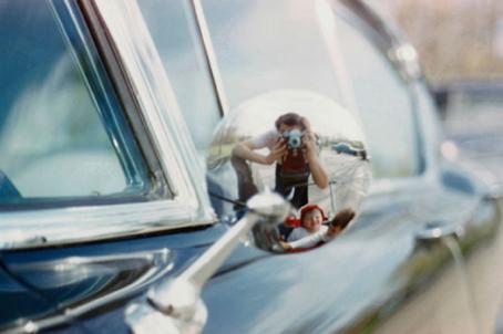 Sinisen auton sivupeilin heijastuksen kautta otettu kuva. Pyöreässä sivupeilissä näkyy nainen kamera nostettuna silmien eteen, sekä kaksi lasta lastenvaunuissa.