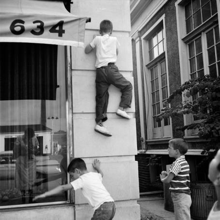Vivian Maier, Chicagoland, ei päiväystä. © Estate of Vivian Maier, Courtesy of Maloof Collection and Howard Greenberg Gallery, NY. Mustavalkoisessa kuvassa kolme pikkupoikaa, joista yksi on kiivennyt seinää pitkin noin puolentoista metrin korkeuteen. Kaksi muuta poikaa seisovat. Kuvaajan hahmo heijastuu seinässä olevasta näyteikkunasta.
