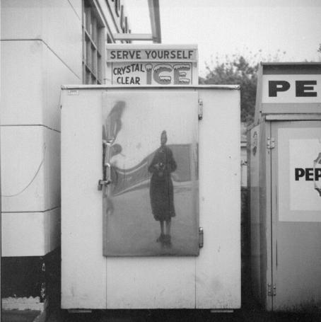Vivian Maier, ei päiväystä. © Estate of Vivian Maier, Courtesy of Maloof Collection and Howard Greenberg Gallery, NY. Mustavalkoinen kuva, jossa kuvaajan hahmo heijastuu aaltoilevana metallisesta pienestä ovesta tai luukusta. Luukku on osa isompaa vaaleaa konttia, jonka päällä on kyltti jossa lukee "Serve yourself crystal clear ice".