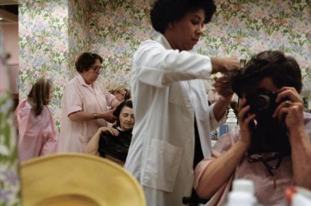 Omakuva, jossa nainen istuu kampaajan tuolissa ja kampaaja käsittelee hänen hiuksiaan. Kamera peittää hänen kasvonsa. Taustalla näkyy muita asiakkaita ja kampaajia. Seinillä on kukkaista tapettia.