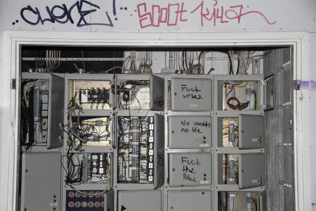Salamalla valaistuja sähkökaappeja. Osa on auki, osa on kiinni ja niissä lukee "Fuck urbex", "No vandalism no life", ja "fuck the police". Yläpuolella seinässä on graffiteja.