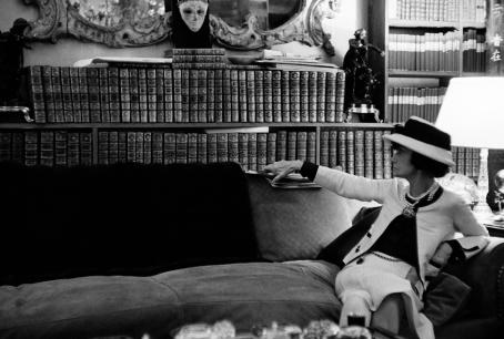 Mustavalkoisessa kuvassa Coco Chanel istuu sohvallaan, hän on kääntyneenä selkänojaa kohti ja ottaa tai laittaa selkänojan päälle. Sohvan takana on kirjahylly täynnä kirjoja, hyllyn päällä on koristeellinen peili tai taulu. 