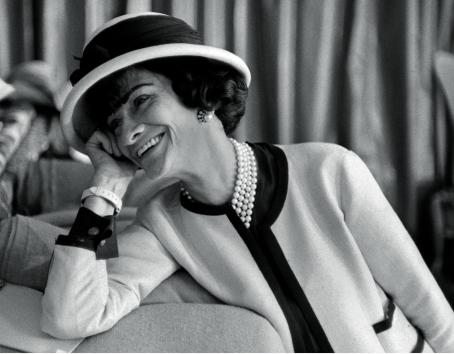 Mustavalkoisessa kuvassa Coco Chanel istuu sohvalla nojaten päätään toiseen käteen. Hän katsoo jotain kuvan ulkopuolella ja hymyilee niin että hänen hampaansa näkyvät. 