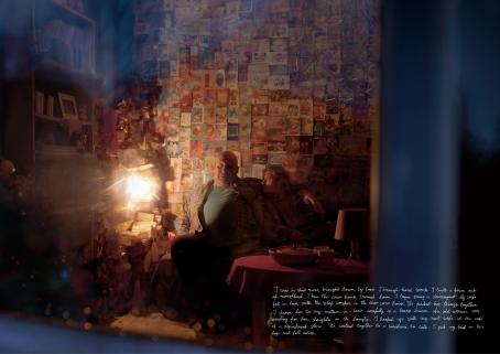 Nainen ja mies istuvat vierekkäin. Mies pitää kättään naisen harteilla, nainen katsoo miestä. Heidän takanaan oleva seinä on kokonaan postikorttien peitossa. Kuvan alareunassa on valkoisella käsin kirjoitettua tekstiä. 