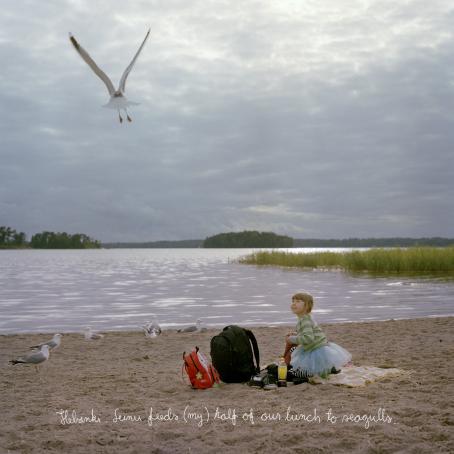 Tyttö istuu hiekkarannalla ja katsoo ylöspäin. Tytön vieressä on isompi ja pienempi reppu. Rannalla on lokkeja, ja yksi lentää ilmassa. Taivas on pilvinen. Alhaalla on käsinkirjoitettu teksti: "Helsinki. Leinu feeds (my) half of our lunch to seagulls."