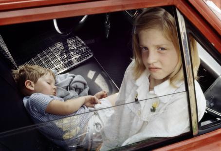 Punaisen auton etupenkillä on kaksi lasta. Isompi tyttö istuu ja katsoo puoliksi auki olevasta ikkunasta kameraan, pienempi poika nukkuu maaten penkillä.