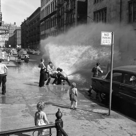 A black and white street view of a city. There is water showering from a fire hydrant on the street. There are many shirtless young boys gathered around it, some of them are either opening it or trying to close it. There are also other people on the street.