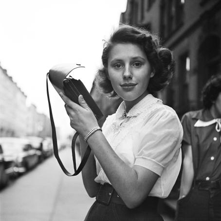 Mustavalkoisessa kuvassa nuori nainen katsoo suoraan kameraan. Nainen pitelee käsissänsä pientä käsilaukkua. Hänellä on lyhyehköt kiharat hiukset, valkoinen paita, iso vyö ja hame tai housut. 