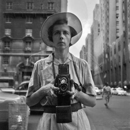 Mustavalkoinen kuva hattupäisen naisen heijastuksesta. Nainen pitää molemmin käsin Rolleiflex-kameraa ja katsoo hieman yläviistoon.