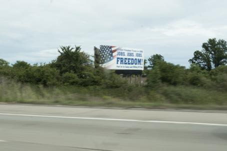 Epäterävä kuva, joka on luultavasti otettu liikkuvasta autosta. Tien varressa kasvillisuuden keskellä on mainos tms. jossa on USA:n lippu ja teksti: Jobs! Jobs! Jobs! FREEDOM!