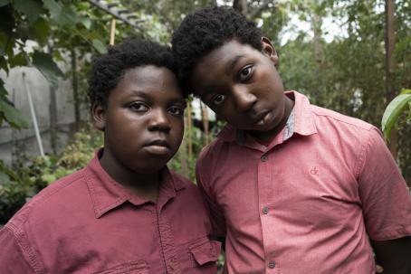 Two black children. Other is a bit taller, and is leaning on the other’s head. Both have a light red collared shirt and both are looking straight into the camera. 