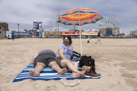 Two white people at the beach on top of a blanket. The woman is sitting, she is wearing a face mask. The man is laying on his back, his face isn’t visible. In the background there are buildings, one says “Tom’s Coney Island” and there is also a ferris wheel. The sun is not shining, the sky is cloudy. 