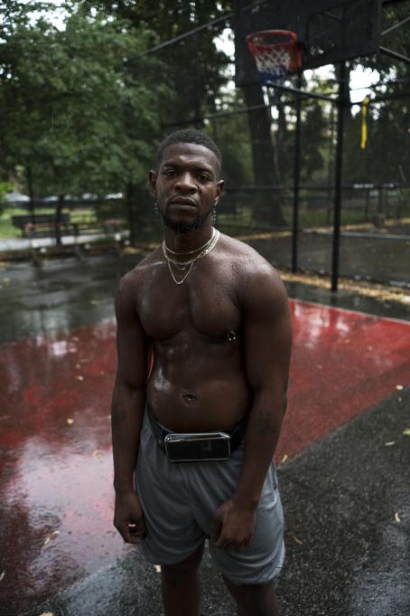Tummaihoinen, paidaton mies seisoo koripallokentällä. Hän katsoo kameraan. Sataa vettä, ja mies on märkä.