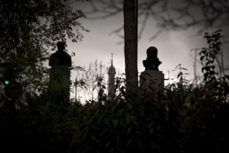 Tummasävyisessä kuvassa näkyy heinikkoa, puita ja taustalla patsaita sekä torni.