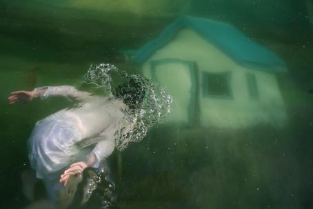 Valkoasuinen hahmo on veden alla. Kuvassa näkyy myös talo. 