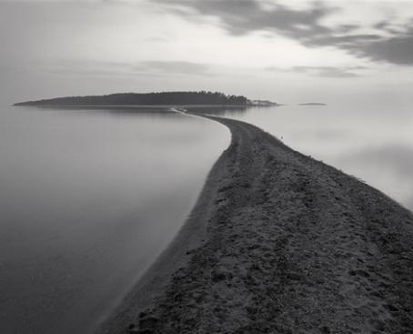 Mustavalkoisessa kuvassa on hiekkasärkkä, joka jatkuu kauempana näkyvää saarta kohti. Särkän ympärillä on tyyni vedenpinta.