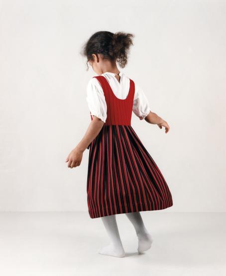 Tummaihoinen tyttö selin päällään valkoinen lyhythihainen paita, valkoiset sukkahousut ja punasävyinen liivimekko päällään.