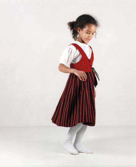 Tummaihoinen tyttö päällään valkoinen lyhythihainen paita, valkoiset sukkahousut ja punasävyinen liivimekko.