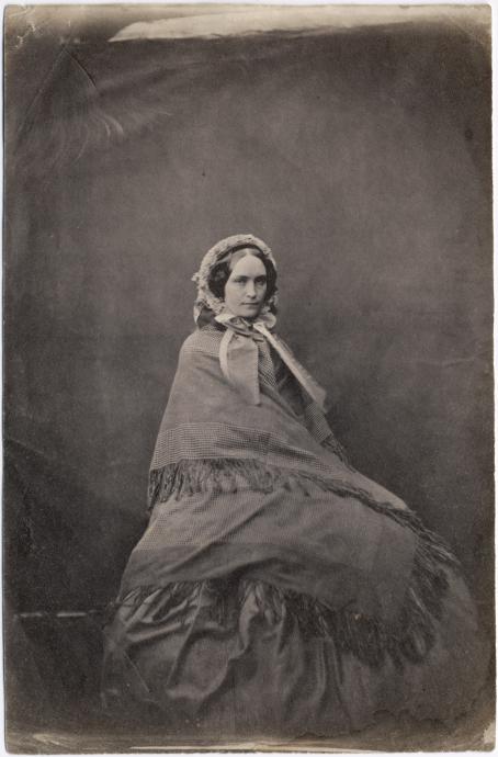 Vanhassa mustavalkoisessa kuvassa istuu nainen, jolla on iso mekko ja hartioilla huivi. Hänellä on myös päässään huppu tai huivi.