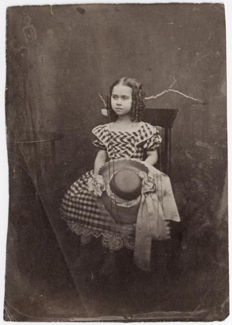 Vanhassa mustavalkoisessa kuvassa tuolilla istuu tyttö, jolla on mekko ja kiharat hiukset. Hän pitää kädessään hattua. 