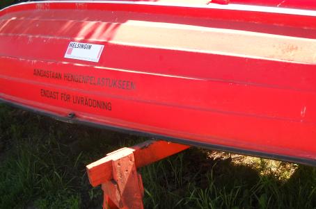 Punainen ylösalaisin oleva vene, jonka alla nurmikkoa. Veneessä lukee "Ainoastaan hengenpelastukseen, endast för livräddning".