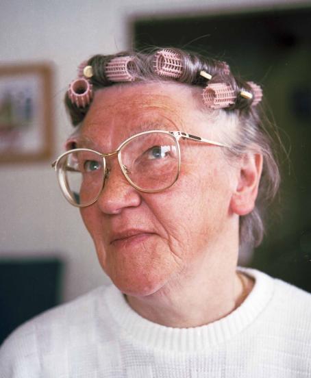 Silmälasipäinen vanha nainen, jolla on papiljotteja hiuksissaan. 