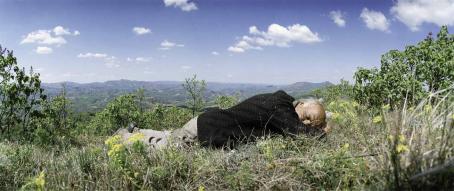 Mies makaa silmät kiinni nurmikolla. Takana näkyy vuoristomaisemaa. 