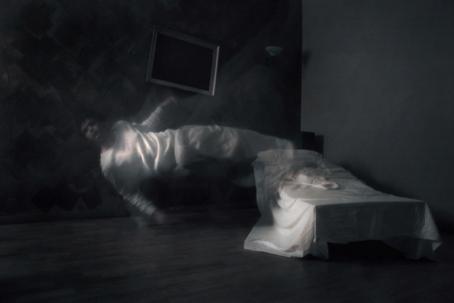 Hämärä huone, jonka reunassa on sänky. Sängyn vieressä on valkoisissa vaatteissa hahmo, joka on ilmassa, vaakasuorassa lattiaan nähden. 