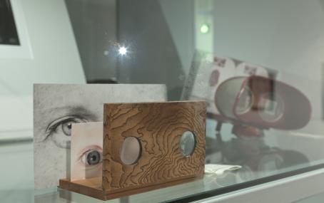 Näyttelyssä oleva teos. Puisessa levyssä on kaksi pientä pyöreää ikkunaa, joiden takana on kaksi kuvaa silmistä. 