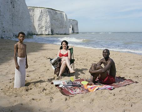 Kolme ihmistä on hiekkarannalla. Tummaihoinen poika seisoo pyyhe lantiolla, valkoihoinen nainen istuu lehti kädessään, tummaihoinen mies istuu pyyhkeen päällä. Heidän takanaan näkyy korkeita valkoisia kallioita.