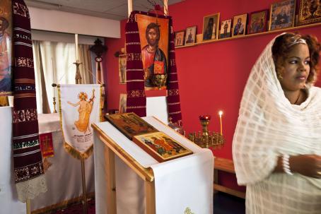 Punaseinäisessä huoneessa on paljon uskonnollisia esineitä ja ikoneita. Oikeassa reunassa on nainen valkoinen huivi päällään.