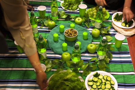 Pöytä johon on katettu kaikkea vihreää. Keskellä on kulhollinen oliiveja, ja sen ympärille on laitettu ympyrään kynttilöitä, omenoita ja mehupulloja. Kaksi kättä laittaa pöydälle lisää tarjottavaa. 