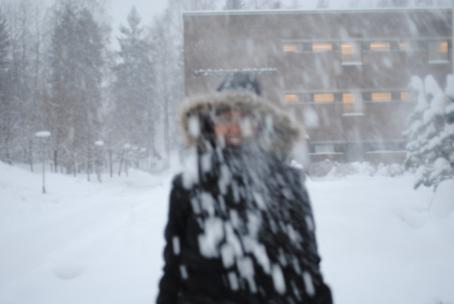 Henkilö musta talvitakki päällään ja huppu päässä, hän näkyy epäselvästi koska sataa lunta. Takana näkyy kerrostalo ja lumista maisemaa. 