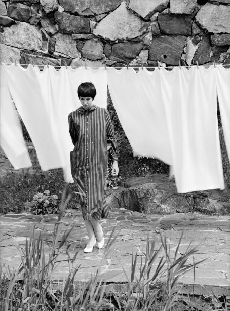 Mustahiuksinen nuori nainen raidallisessa mekossa seisoo pyykkinarulla roikkuvien valkoisten lakanoiden edessä. 