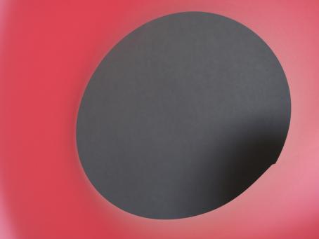 Vaaleanpunaisella taustalla harmaansävyinen ympyrä, jonka yhdellä reunalla harmaa tummenee mustaksi.